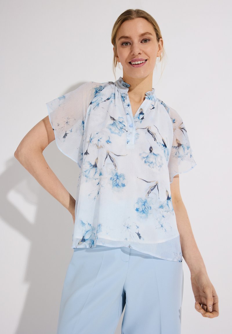Young woman smiling, wearing a sheer white blouse with blue floral print and light blue pants, standing against a plain white background.