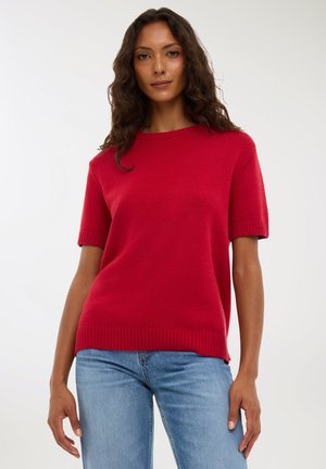 Maglione rosso lavorato a maglia con maniche corte, polsini e orlo a coste. Abbinato a jeans azzurri chiari, che mostrano una vestibilità rilassata e una texture morbida.