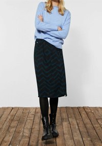 Černá sukně se zeleným zigzag vzorem, kombinována se světle modrým svetrem a černými punčochami, doplněná černými kotníkovými botami. Pozadí tvoří dřevěná podlaha.