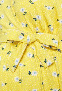 Tecido amarelo com pequenas flores brancas e detalhes azuis. Apresenta uma cintura ajustada com um detalhe de laço e um padrão texturizado com pontos.