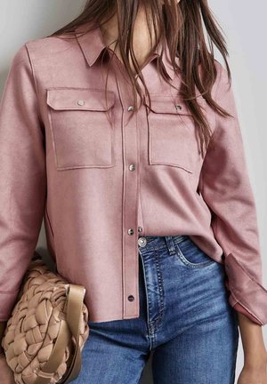 Vrouw draagt een roze overhemd met knopen en klepzakken en blauwe spijkerbroek, houdt een geweven beige handtas aan haar zijde.