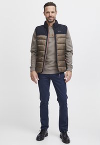 Gepolsterte Weste in Dunkelbraun und Marineblau, kurzer Reißverschluss, kombiniert mit einem hellgrauen Sweatshirt, Marineblauen Hosen und schwarzen Schnürschuhen.