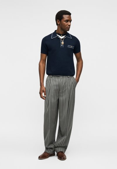 Homme portant un polo bleu marine à manches courtes avec un liseré blanc, un pantalon large gris à rayures verticales, des chaussures marron et des colliers en or superposés, debout les mains dans les poches.