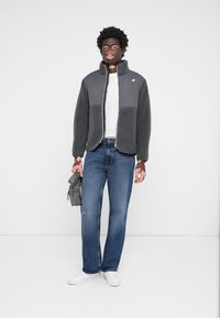 Veste en polaire grise avec un col haut, fermeture éclair sur le devant, design bicolore, portée sur une chemise blanche et associée à un jean bleu et des baskets blanches.