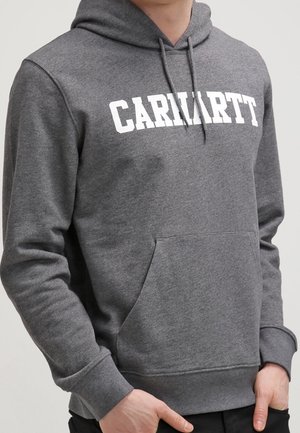 Osoba nosząca szarą bluzę Carhartt z kapturem, sznurkami i kieszenią z przodu, ręce swobodnie spoczywają w pobliżu kieszeni.