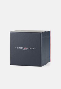Boîte noire avec "MONTRES TOMMY HILFIGER" en texte blanc. La boîte présente des accents rouges et bleus sur les côtés. Texture lisse, fermée avec un couvercle.