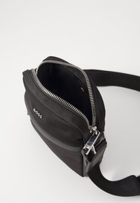 Borsa a tracolla nera realizzata in tessuto testurizzato, con una zip argentata e una tracolla regolabile. Nella parte anteriore c'è una piccola tasca con un logo.