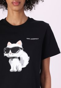 Tricou negru din bumbac cu un pisoi de desen animat alb purtând ochelari de soare, evidențiat de textul "KARL LAGERFELD" în alb. Mâneci scurte, guler rotund.