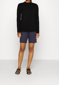 Schwarzes Langarmshirt, marineblaue Shorts und schwarze Sandalen. Das Oberteil hat einen runden Halsausschnitt; die Shorts sind knielang mit Umschlägen.