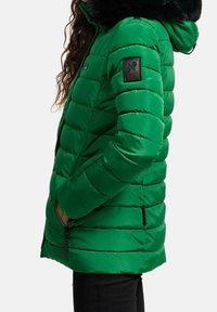 Chaqueta acolchada verde con capucha de pelo sintético negro, bolsillos con cremallera y un diseño acolchado con un parche de logo en la manga izquierda.