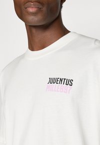Camiseta de algodão branca com um design impresso, com "JUVENTUS" em preto e "MILLE897" em rosa. Decote redondo, mangas curtas.