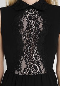 Robe noire avec un insert en dentelle à motifs floraux, un col festonné et un corsage fluide, créant une texture contrastée.