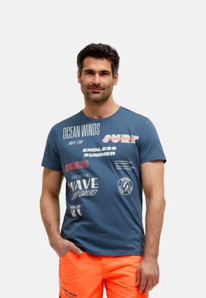 Mann trägt blaues Grafik-T-Shirt und leuchtend orangefarbene Shorts, steht mit einer Hand in der Tasche vor einfachem Hintergrund.