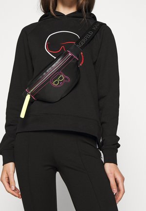 Persona che indossa un hoodie nero e pantaloni, con una borsa a tracolla nera accentata al neon che presenta un logo stilizzato di un volto con occhiali da sole.