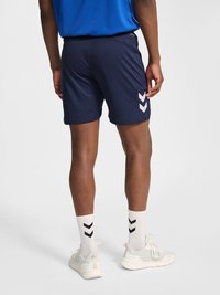 Osoba u mornarskoplavim sportskim kratkim hlačama s bijelim logoima u obliku cik-cak crte, bijelim čarapama do gležnja s crnim cik-cak crtama i bijelima tenisicama, stoji na sivom podu.