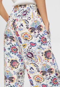 Mains dans les poches d'un pantalon blanc orné de motifs floraux cachemire colorés, porté avec un haut blanc côtelé.