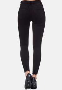 Jean skinny noir extensible taille haute, avec deux poches arrière et ourlets effilochés. Porté avec des chaussures à talons.