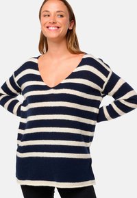 Femme souriante les mains derrière le dos, portant un pull V en rayures bleu marine et blanches et un pantalon noir, sur fond uni.