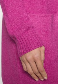 Fuchsia Pullovermanschette mit geripptem Muster, aus weichem, gestricktem Material. Nahaufnahme einer Hand, die auf dem Ärmel ruht.