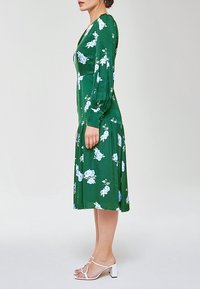 Femme de profil portant une robe verte à manches longues, longueur genou, à fleurs blanches et des sandales blanches à talons, debout devant un fond uni.