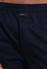 Tummansinisiä puuvillahousuja, joissa on joustava vyötärö, ja takana pieni sininen kangastunnus, jossa lukee "AMMANN". Pehmeä materiaali.