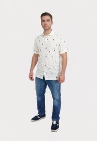 Kortærmet hvid polo skjorte med blå sejlbåd og geometriske mønstre, knapkrave, parret med blå jeans og mørke sneakers.