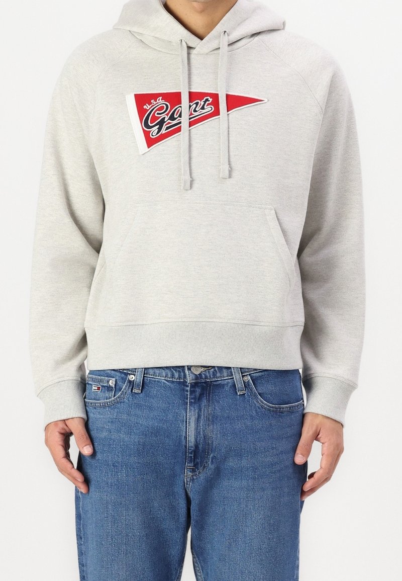 Sweat à capuche gris clair avec poche avant et écusson triangulaire rouge portant le logo "USA Gant", porté avec un jean bleu par une personne debout.