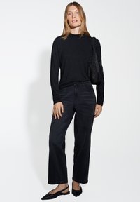 Schwarzes Langarmshirt mit dezentem Glitzer und rüschigem Ausschnitt, kombiniert mit hochgeschnittenen schwarzen weitbeinigen Jeans und schwarzen Slingback-Schuhen mit spitzer Zehenpartie.
