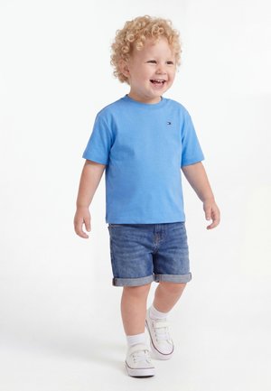 Μικρό παιδί με σγουρά μαλλιά που χαμογελάει, φορώντας μπλε μπλουζάκι, τζιν σορτς, λευκές κάλτσες και λευκά αθλητικά παπούτσια, στέκεται σε λευκό φόντο.