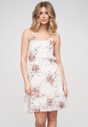 Ung kvinde med blondt hår iført en hvid blomsterprintet, ærmeløs kjole med flæsekant, stående mod en ensfarvet baggrund.