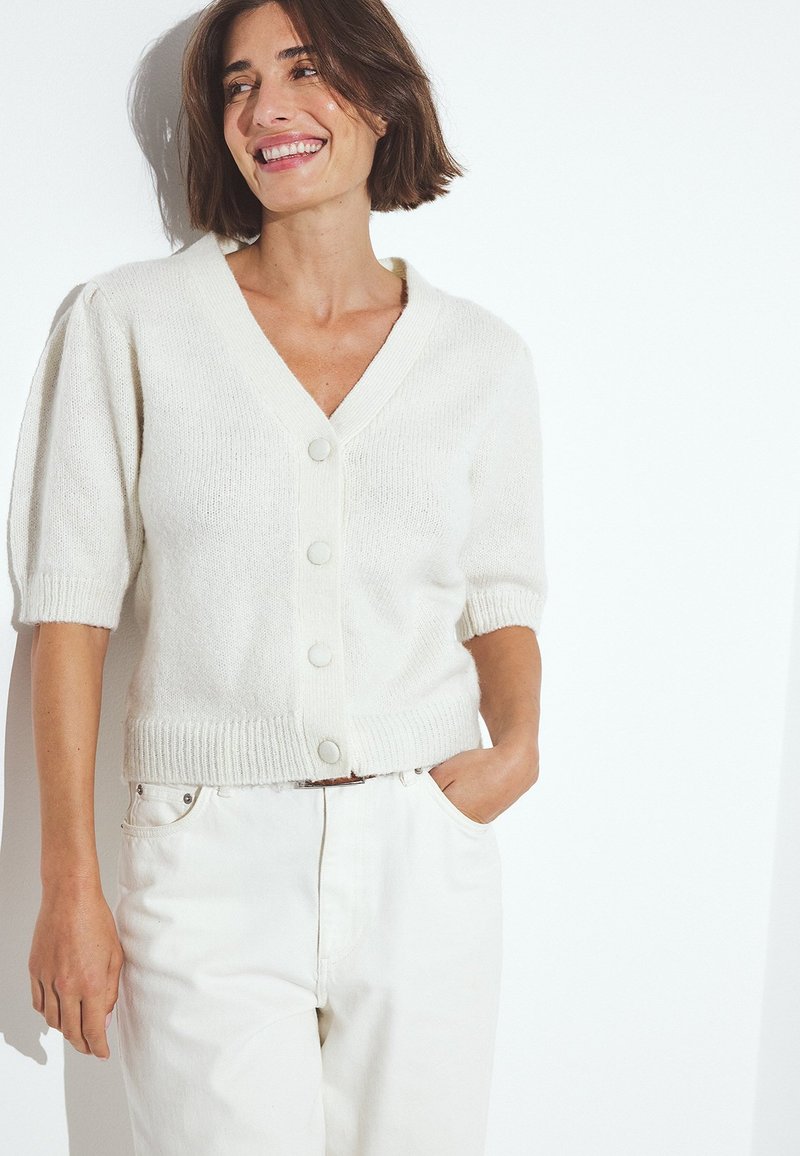 Vit stickad cardigan med korta puffärmar, V-ringning och sex framknappar. Bärs över ljusa byxor med strukturerad yta.