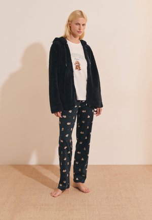 Mujer con chaqueta negra peluda, camiseta blanca con gráfico de perro y pantalones de pijama negros con pequeños dibujos de perros, de pie descalza sobre suelo beige.
