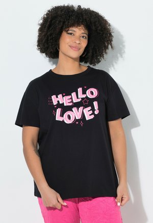 Frau mit lockigem Haar, die ein schwarzes T-Shirt mit pinkfarbenem „HELLO LOVE!“-Schriftzug und leuchtend pinke Hosen trägt, steht vor einem weißen Hintergrund.