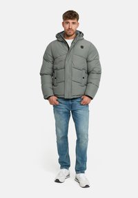 Homme debout avec les mains dans les poches, portant une doudoune grise, un jean bleu et des baskets blanches sur fond blanc.