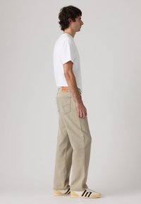 Φωτεινό μπεζ τζιν με φαρδιά γραμμή, συνδυασμένο με λευκή μπλούζα και κρεμ αθλητικά παπούτσια με μαύρες ρίγες. Ξεχωριστή κόκκινη ετικέτα της μάρκας στο πίσω μέρος.