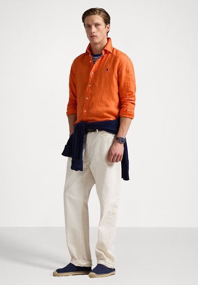 Cămașă portocalie din in cu nasturi, tricou în dungi pe dedesubt, pantaloni albi, pulover bleumarin legat la talie, espadrile albastre, look casual.