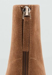 Primo piano di un stivale in suede marrone che mostra una zip verticale con un tassello in suede e cuciture dettagliate.