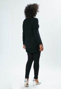 Schwarze Tunika mit langen Ärmeln und seitlichen Schlitzen, kombiniert mit schwarzen Leggings. Das Outfit wird durch silberne High Heels ergänzt.