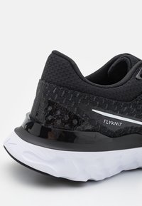 Scarpa sportiva nera con tomaia Flyknit testurizzata, tallone nero lucido, dettagli bianchi e suola bianca ammortizzata con un design ondulato.