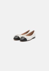 Ballerines blanches avec des bouts ronds noirs et de petits nœuds sur chaque chaussure, sur un fond blanc uni.