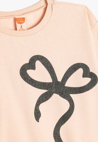 Felpa rosa chiaro con una grande grafica a fiocco grigio scuro. Il tessuto sembra morbido, con colletto a coste e maniche corte.