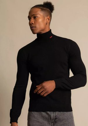Svart polotröja i ribbat material med långa ärmar. Har en liten röd logotyp vid halsen. Kombinationen bärs med grå jeans.