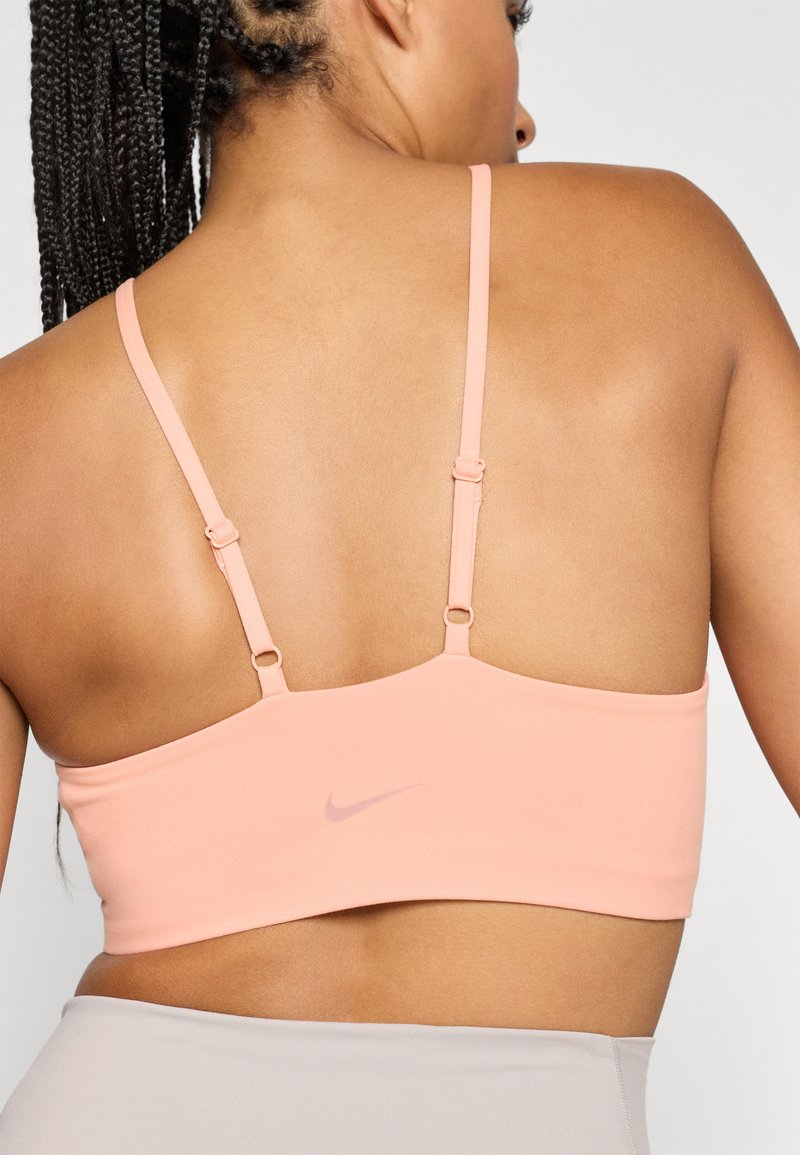 Pfirsichfarbener Sport-BH mit dünnen verstellbaren Trägern und einem Verschluss auf der Rückseite. Verfügt über ein dezentes Logo in der Mitte und eine glatte Stoffoberfläche.