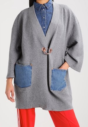 Mujer con un cárdigan gris texturizado con grandes bolsillos azules de mezclilla y un broche decorativo, sobre una camisa de mezclilla azul y pantalones rojos.