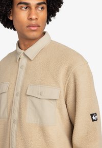 Giacca camicia in pile beige con due tasche sul petto, bottoni a pressione e collo a contrasto. Patches con logo sulla manica. Tessuto morbido e testurizzato.