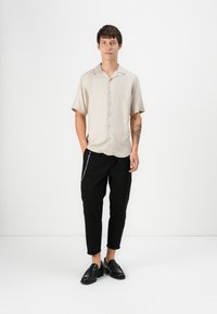 Short-sleeve beige shirt with button-down front, paired with black pants featuring a silver chain on the side, and black loafers.