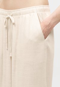 Φωτεινό μπεζ παντελόνι από λινό με ελαστική μέση και κορδόνι, πλαϊνή τσέπη με το χέρι εν μέρει μέσα.