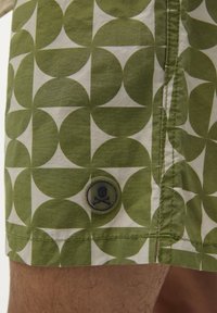 Șorturi de baie cu un model verde și alb, având cercuri suprapuse, un buton verde solid cu un design de skull și detalii de cusături vizibile.
