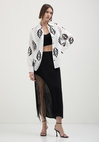 Camicia bianca con motivi a foglia nera, abbinata a un top corto nero e una gonna lunga con dettagli in frange. Tacchi neri con laccetti.