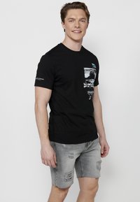 Juodas medvilninis marškinėlis su pilkų atspalvių grafika priekyje, apvaliu kaklu ir trumpomis rankovėmis, derinamas su nusidėvėjusiais pilkais džinsiniais šortais.